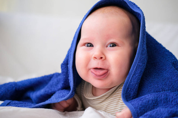 Cute smiling caucasian infant baby looking at camera in blue towel after bathing in the bath on white bed.Copyspace. - Foto, Imagem