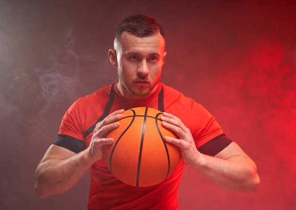 Nuori urheilullinen koripalloilija hyökkäysasennossa pitäen palloa kahdella kädellä oranssilla ja punaisella taustalla savulla
 - Valokuva, kuva