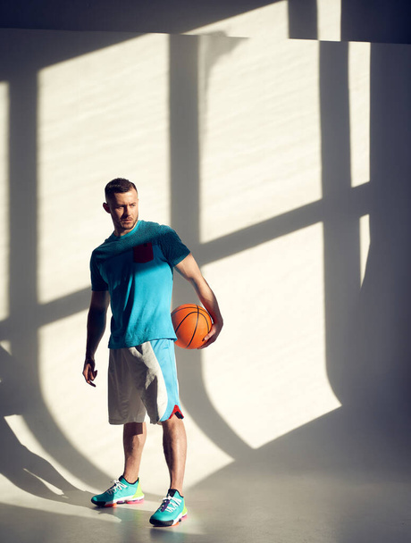 Młody sportowiec, koszykarz trzymający piłkę i stojący przy ścianie z cieniami z okna - Zdjęcie, obraz