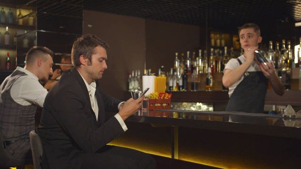 Mies istuu puhelimessa baarissa, baarimikko ravistelee alkoholia.
 - Valokuva, kuva