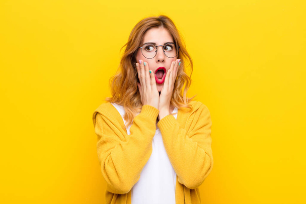 young pretty blonde woman feeling shocked and scared, looking terrified with open mouth and hands on cheeks against yellow wall - Foto, afbeelding