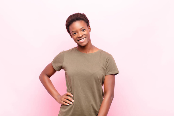 young pretty black womansmiling happily with a hand on hip and confident, positive, proud and friendly attitude against pink wall - Foto, Bild