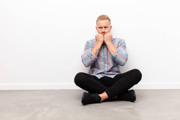 young blonde man looking worried, anxious, stressed and afraid, biting fingernails and looking to lateral copy space sitting on cement floor - Фото, изображение