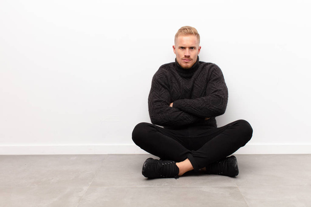 young blonde man feeling displeased and disappointed, looking serious, annoyed and angry with crossed arms sitting on cement floor - Фото, зображення