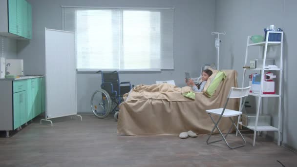 Tyttö makaa sängyllä sairaalassa, pitää tabletin käsissä ja katso jonnekin
 - Materiaali, video