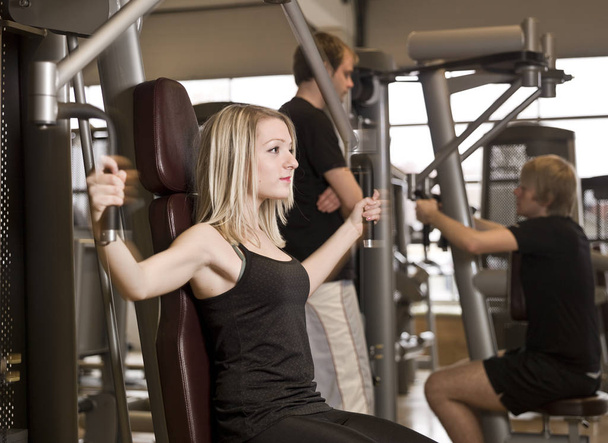 Girl using an exercise machine at a health club with two men in the background - 写真・画像