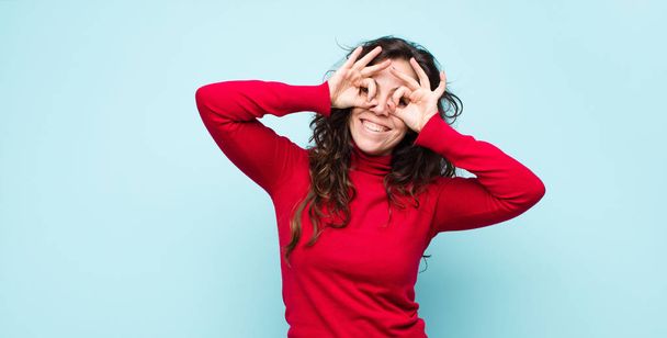 young pretty woman feeling shocked, amazed and surprised, holding glasses with astonished, disbelieving look against blue wall - Fotoğraf, Görsel