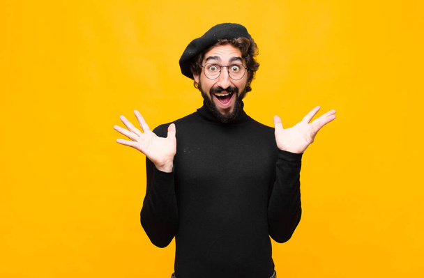young french artist man looking happy and excited, shocked with an unexpected surprise with both hands open next to face against orange wall - Foto, Bild