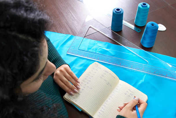 μοδίστρα γράφει μέτρα σε ένα σημειωματάριο. ο ράφτης δημιουργεί μια συλλογή από ρούχα. νεαρή γυναίκα σχεδιαστής κάνει σημειώσεις των ιδεών στο σημειωματάριο - Φωτογραφία, εικόνα