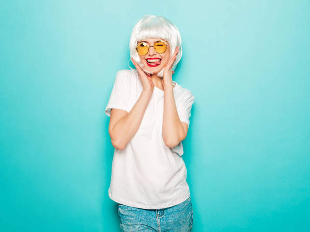 Young sexy smiling hipster girl in white wig and red lips.Beautiful trendy woman in summer clothes.Carefree model posing near blue wall in studio.Positive female going crazy - Foto, Bild