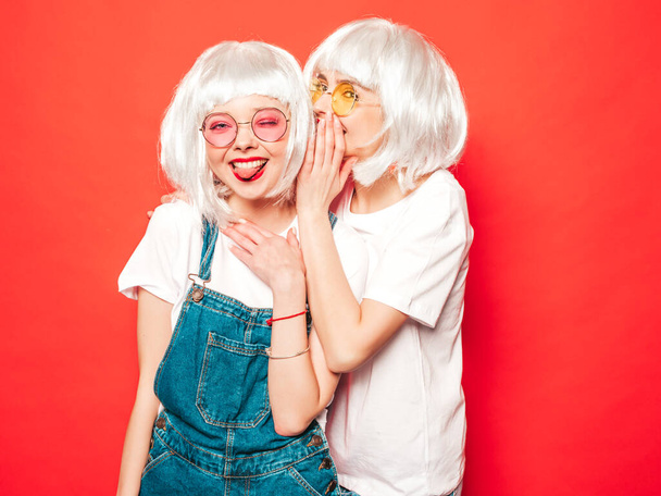 Two young sexy hipster girls in white wigs and red lips.Beautiful trendy women in summer clothes.Carefree models posing near red wall in studio.Positive female shares secret,gossip - Fotó, kép