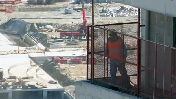 Engineer watching the construction site. A worker looking down from the balcony. Work safety. Helmet, hard hat, vest. Red orange iron fence. High skyscraper constructions. Single one solo waiting 4K. - Footage, Video