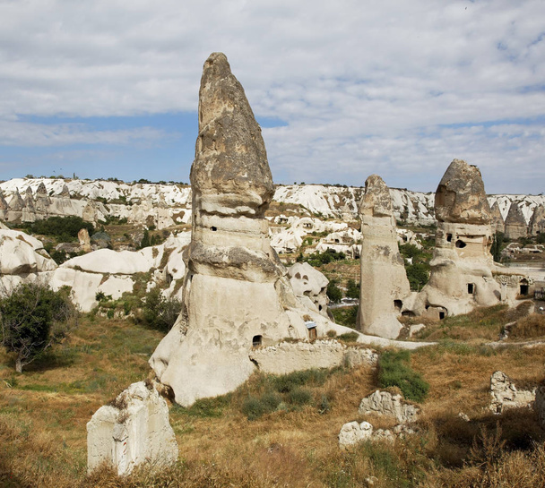 square format group of limestone fairy chimney homes near Goreme Cappadocia Turkey, volcanic lava hills in the background, cloudy blue sky with crop space and copy space - Fotó, kép