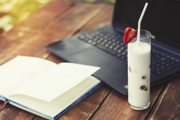 Laptop, otevřený deník a sklenici s mlékem osvěžující nápoj s jahodami na dřevěný stůl a v pozadí na zahradě. Koncepce práce v přírodě, volné noze, pracujte na dovolené - Fotografie, Obrázek