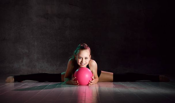 Young smiling girl gymnast in black sport body and uppers sitting in twine on floor and holding pink gymnastic ball in hands - Fotografie, Obrázek