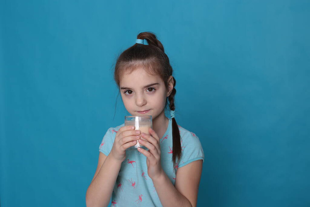 Beautiful happy girl of eight years old drinks warm milk from a transparent glass on a blue background in a blue T-shirt in a healthy state - 写真・画像