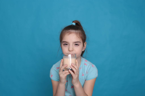 Beautiful happy girl of eight years old drinks warm milk from a transparent glass on a blue background in a blue T-shirt in a healthy state - Fotoğraf, Görsel