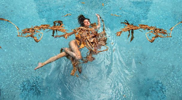 Top view of a beautiful young sexy woman floating elegantly relaxed in dried up leaves of a banana tree, turquoise water in the pool, copy space - Photo, Image