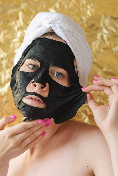 Young woman apply black cosmetic fabric facial mask on golden background. Face peeling mask with charcoal, spa beauty treatment, skincare, cosmetology. Close up - Foto, Imagem