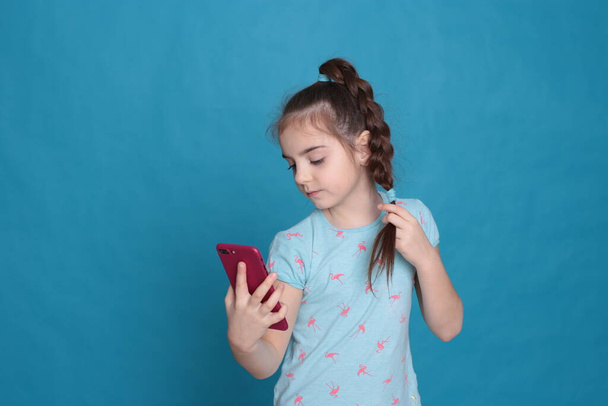 Ένα χαμογελαστό κορίτσι οκτώ ετών κρατά ένα ροζ τηλέφωνο στα χέρια της σε ένα μπλε φόντο - Φωτογραφία, εικόνα