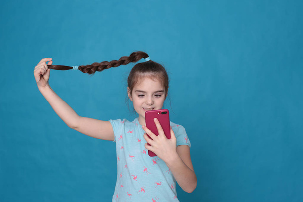 A smiling girl of eight years old holds a pink telephone in her hands on a blue background - Foto, Bild