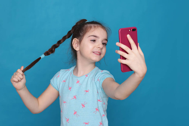 A smiling girl of eight years old holds a pink telephone in her hands on a blue background - Photo, Image