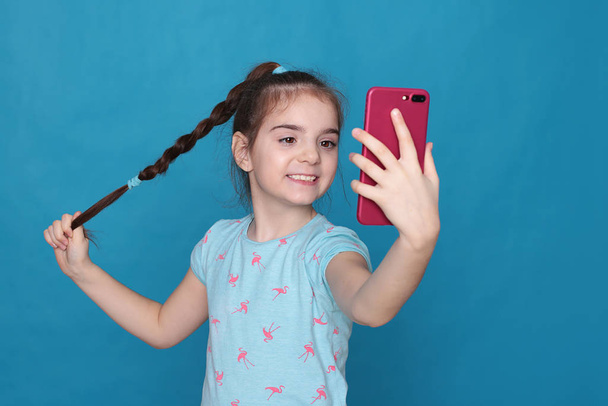 A smiling girl of eight years old holds a pink telephone in her hands on a blue background - Photo, Image