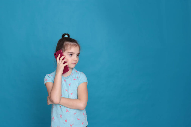 Egy nyolc éves mosolygós lány rózsaszín telefont tart a kezében, kék alapon. - Fotó, kép