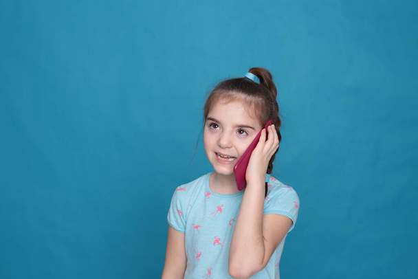 A smiling girl of eight years old holds a pink telephone in her hands on a blue background - Fotografie, Obrázek