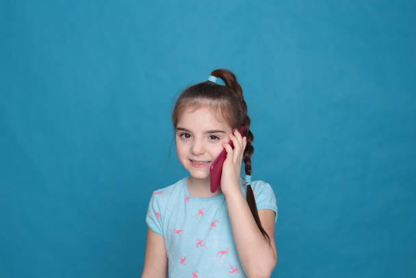 Egy nyolc éves mosolygós lány rózsaszín telefont tart a kezében, kék alapon. - Fotó, kép
