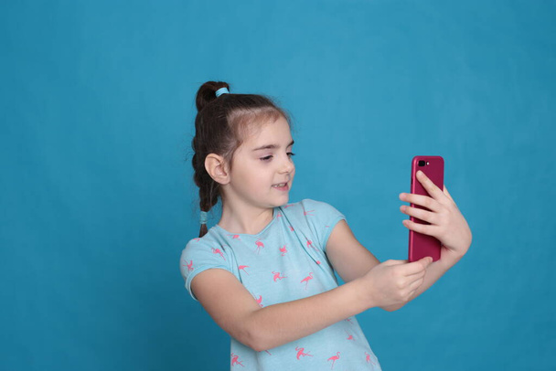 Uma menina sorridente de oito anos segura um telefone rosa em suas mãos em um fundo azul
 - Foto, Imagem