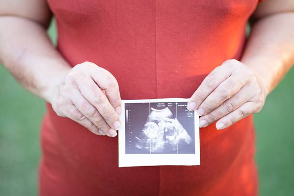 Detailní záběr těhotné ženy v červených mateřských šatech drží ultrazvuk obrázek rostoucího dítěte na její dítě hrbol. - Fotografie, Obrázek