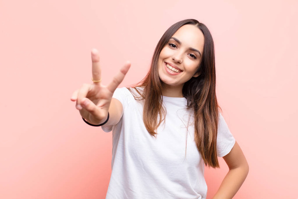 young pretty woman smiling and looking happy, carefree and positive, gesturing victory or peace with one hand against pink wall - Fotoğraf, Görsel