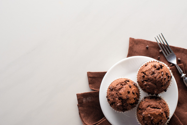 top view of fresh chocolate muffins on white plate and brown napkin near fork on marble surface - Photo, Image