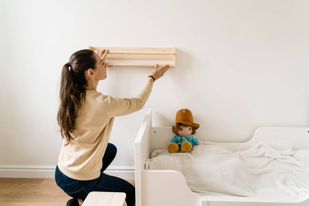 Young woman renovation child room in new house. Wooden bookshelf. Neutral nursery - Фото, изображение