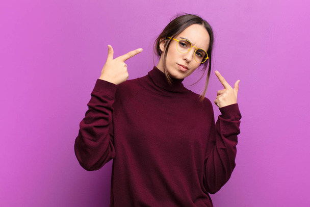 young pretty woman with a bad attitude looking proud and aggressive, pointing upwards or making fun sign with hands against purple wall - Fotó, kép