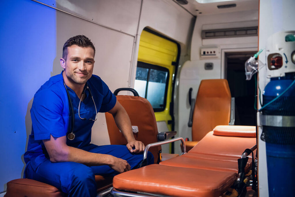 El forense de uniforme se sienta en el coche de la ambulancia y sonríe
 - Foto, Imagen