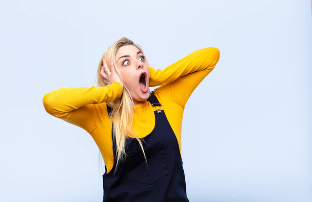 young pretty blonde woman with open mouth, looking horrified and shocked because of a terrible mistake, raising hands to head against flat wall - Фото, зображення