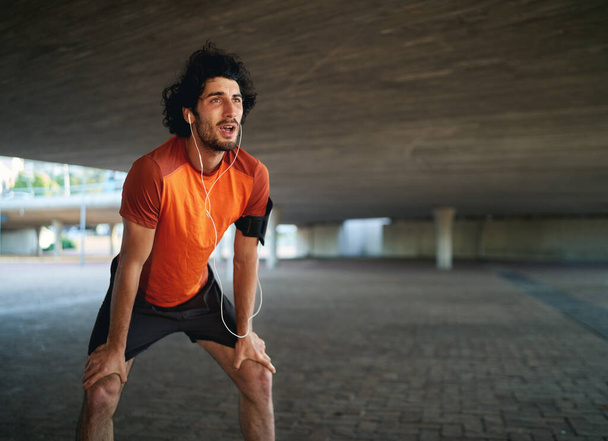 Портрет уставшего кавказского спортсмена, слушающего музыку, дышащего после долгой пробежки под мостом
 - Фото, изображение