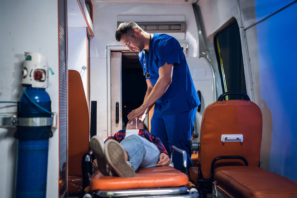 Dokter in medisch uniform zet zuurstofmasker op vrouw liggend op brancard in de ambulance auto - Foto, afbeelding