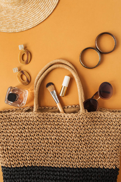 Straw hat, rattan beach bag with women's accessories (sunglasses, earrings, perfume, bracelet, lipstick, brush) inside on ginger background. Flatlay, overhead view. Modern fashion blog collage. - Photo, Image