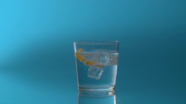 Feche o gelo e uma fatia de limão girando em um copo com água mineral espumante em um fundo azul. Água mineral naturalmente carbonatada
 - Filmagem, Vídeo