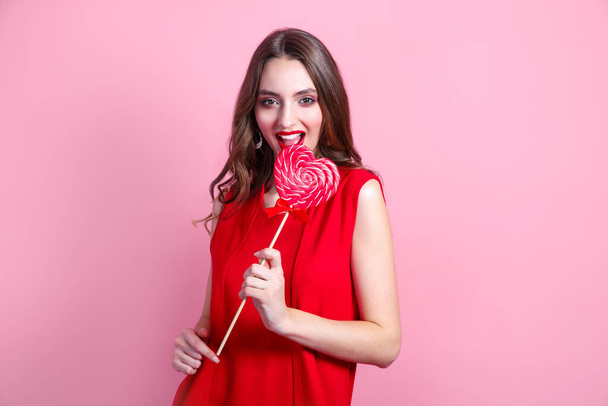 Beautiful brunette girl holding a red heart-shaped lollipop on pink background. Valentines Day - Foto, afbeelding