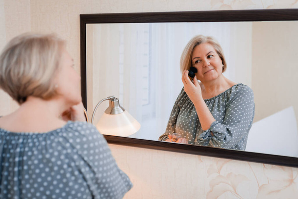 happy middle aged elderly woman preens and adjusts her hair in front of the mirror of the dressing table - Foto, afbeelding