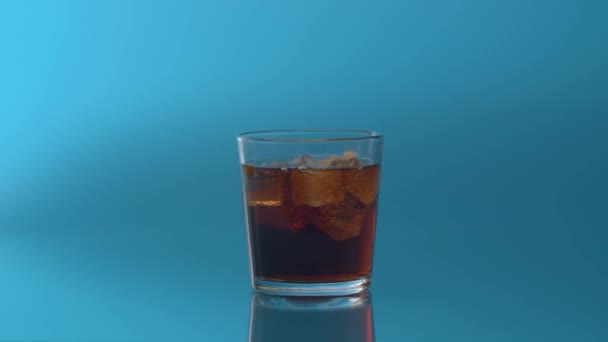 Een glas cola met ijs en citroen van dichtbij. Cola frisdrank met ijsblokjes en gesneden citroen draaiend in doorzichtig drinkglas over blauwe achtergrond. Cola Soda met belletjes close-up. - Video