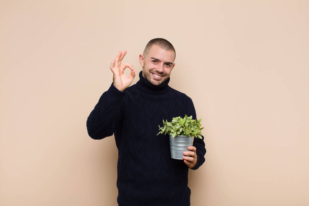 young handsome man feeling happy, relaxed and satisfied, showing approval with okay gesture, smiling holding a plant - Foto, Imagen