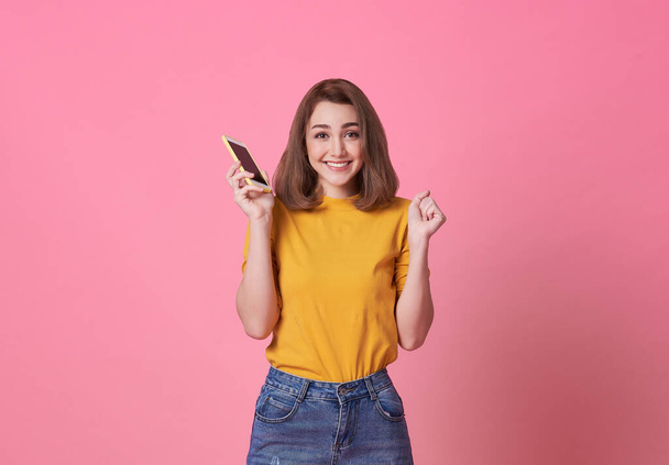 Πορτρέτο της ευτυχούς νεαρής γυναίκας που κρατά το κινητό τηλέφωνο απομονωμένο σε ροζ φόντο. - Φωτογραφία, εικόνα