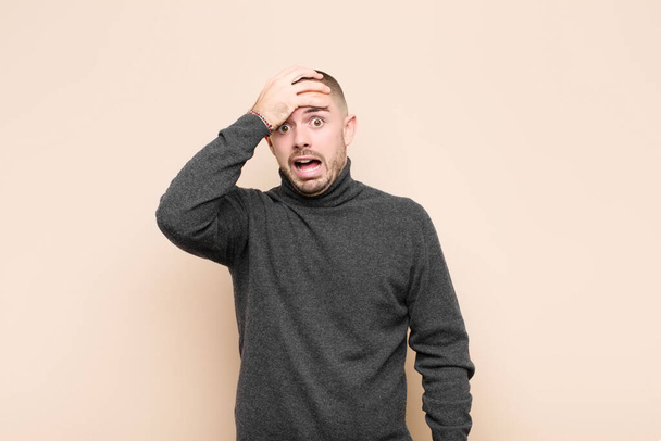 young handsome man panicking over a forgotten deadline, feeling stressed, having to cover up a mess or mistake against flat wall - Fotoğraf, Görsel