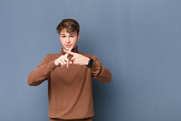 young blonde man looking serious and displeased with both fingers crossed up front in rejection, asking for silence isolated against flat wall - Foto, imagen