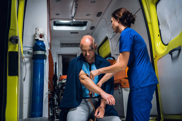 Hübsche Krankenschwester in Uniform wickelt im Krankenwagen ein Tourniquet um die Hand eines Mannes in Decke - Foto, Bild